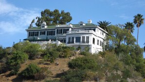 Une grande maison à deux étages, dotée d’un toit végétalisé, entourée d’une végétation luxuriante et de palmiers.