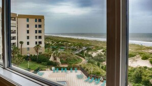 A view of a beach resort with a pool area, palm trees, and a building in the background. - Surf & Racquet Club C126 - Pool! (Amelia Island)