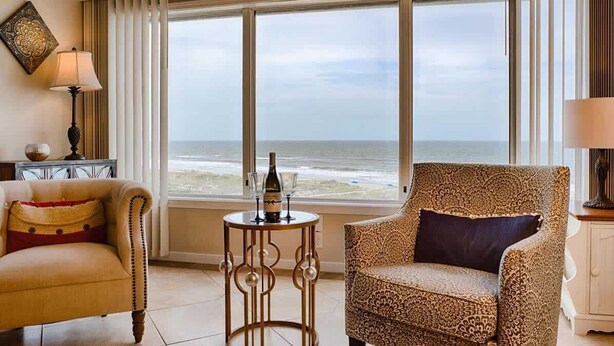 A room with a view of the ocean, featuring two patterned armchairs, a small round table with a bottle and glasses, and a lamp on a side table. - Surf & Racquet Club C126 - Pool! (Amelia Island)