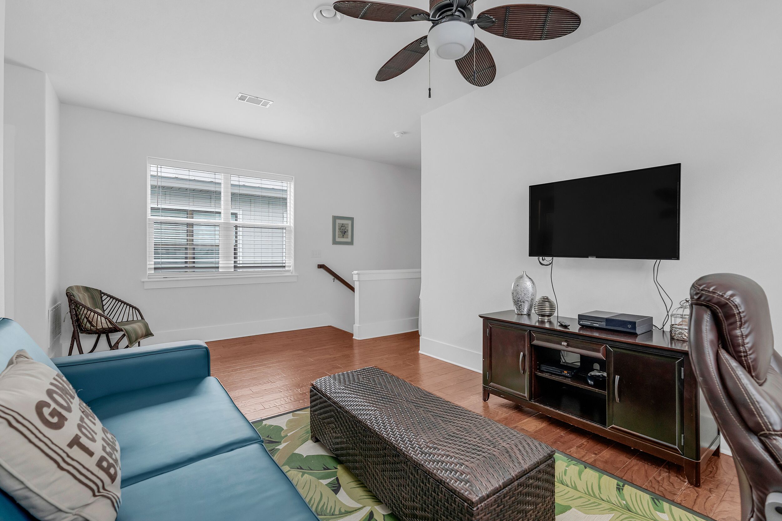 A living room with a blue sofa, a wicker bench, a dark wooden entertainment unit, a flat-screen TV, and a ceiling fan.