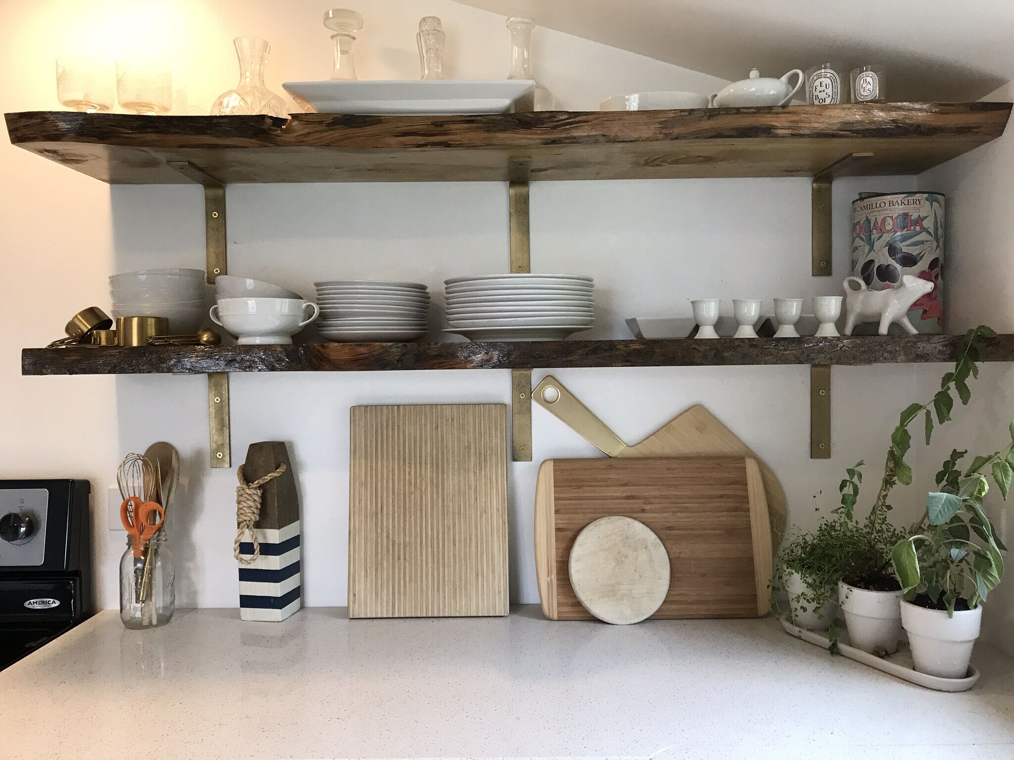 Une cuisine avec des étagères en bois où sont rangées des assiettes, des bols et des bocaux. On y trouve également une planche à découper, une plante et un porte-épices.