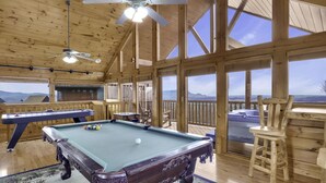 A wooden interior with a pool table, ceiling fans, and large windows offering a mountain view.
