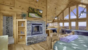 A cozy cabin interior with a stone fireplace, wooden walls, a pool table, and a large window offering a scenic view.