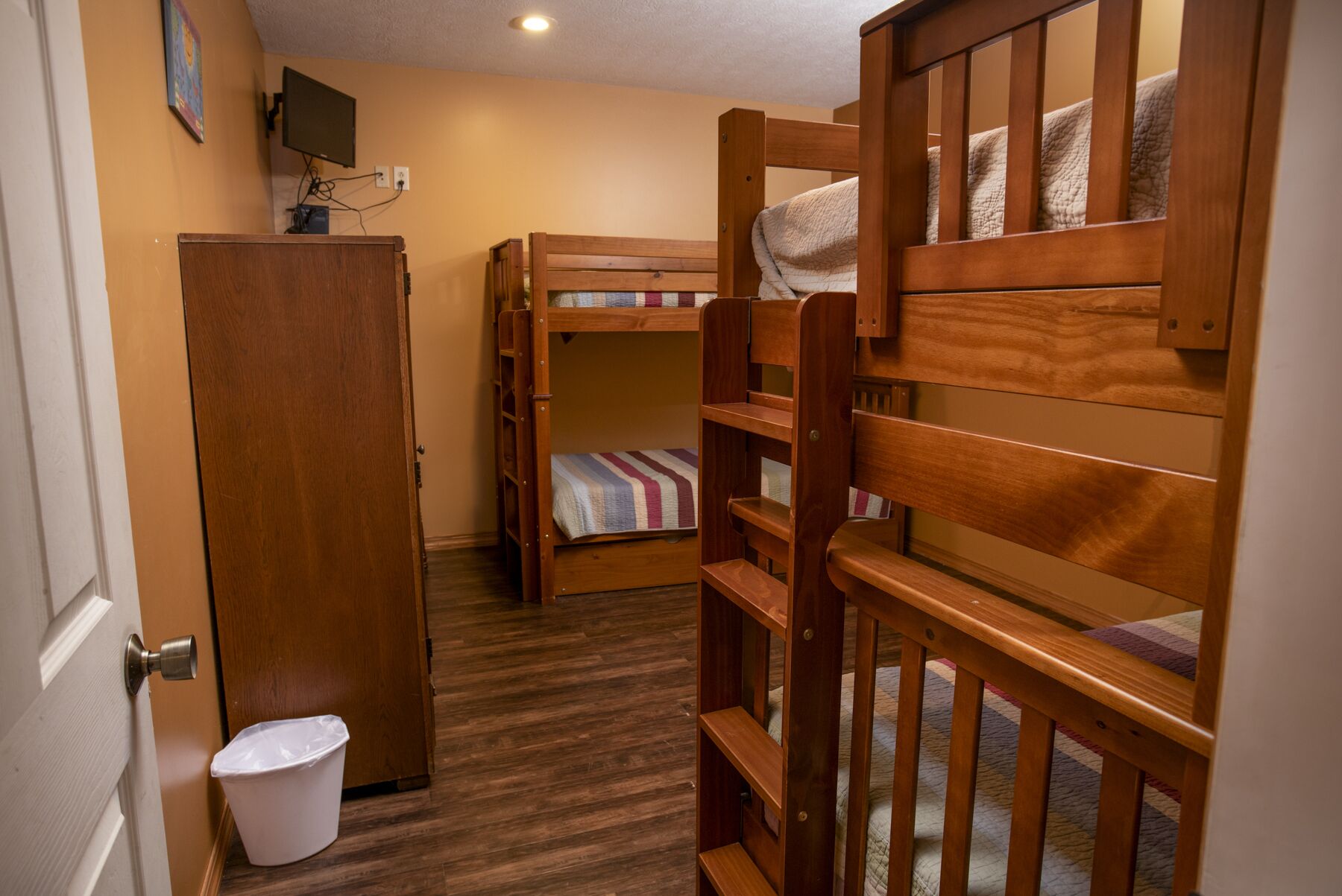 Ein Schlafsaalzimmer mit Etagenbetten, einem Holzschrank, einem an der Wand befestigten Fernseher und einer weißen Mülltonne.