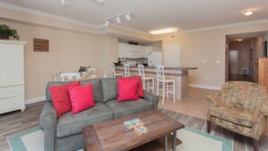 A living room with a grey sofa, red cushions, a wooden coffee table, and a floral armchair. There is a kitchen in the background with white cabinets and a microwave.