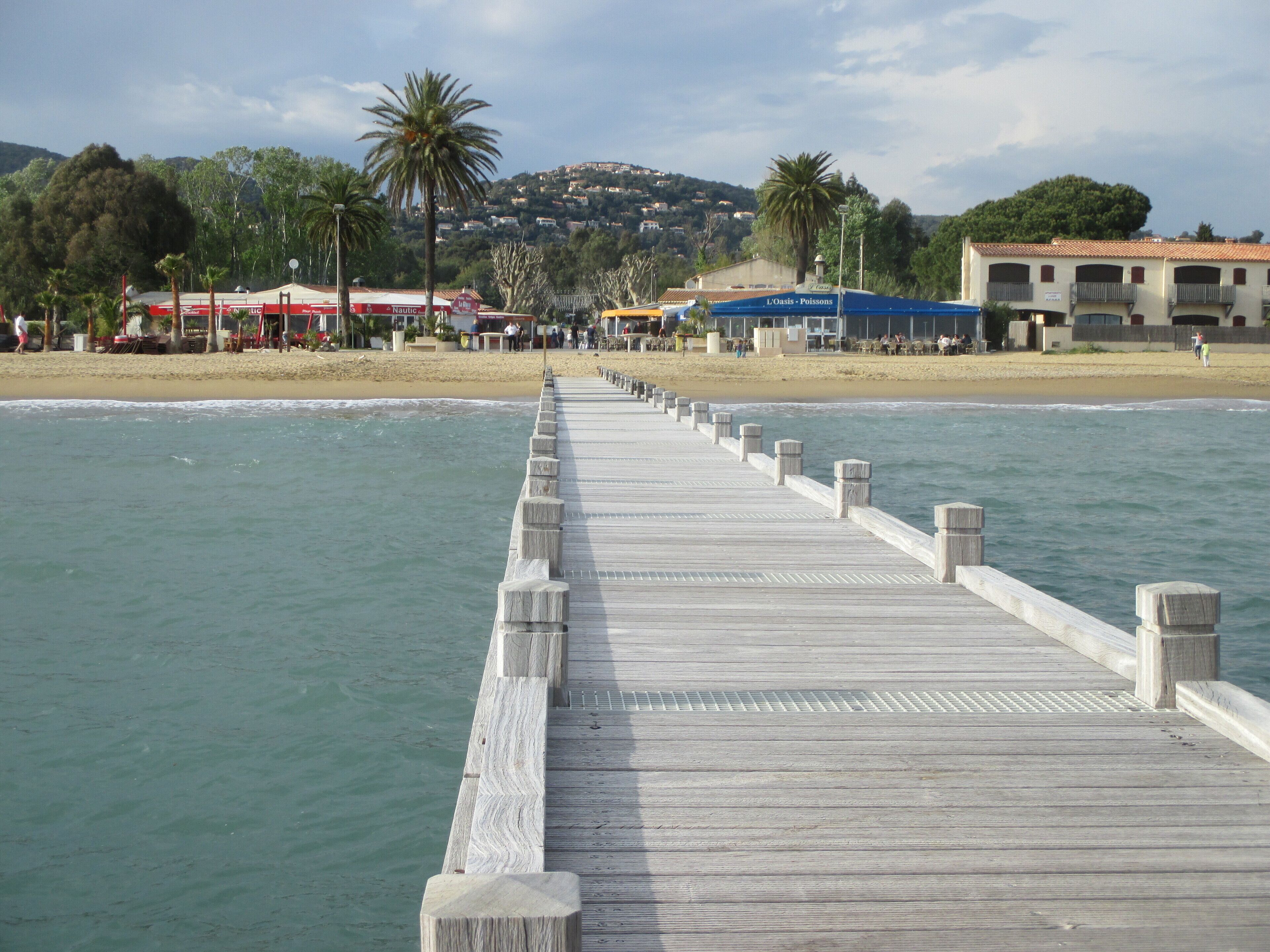 A wooden pier extending into calm waters with a beach and buildings in the background.