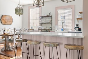 Eine Küche mit einer weißen Kochinsel, Barhocker, einem Kronleuchter und einem Holzschneidebrett an der Wand.