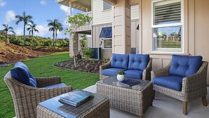 Un patio con mobiliario de mimbre y una mesa de vidrio, con vistas a un jardín con palmeras y una casa al fondo.