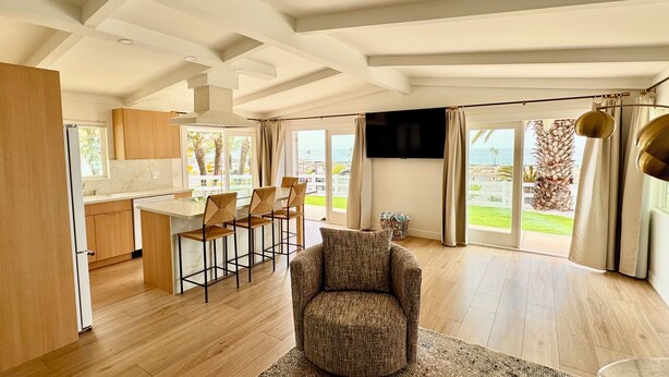 A spacious living area with a large window, a comfortable armchair, and a kitchen with wooden cabinets and a central island. - Best Spot in Carlsbad! Private Yard, Ocean View, Walk to beach.  (Carlsbad)