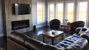 A spacious living room with a fireplace, leather sofas, and large windows offering a view of the outdoors. - Picturesque oceanfront / beachfront estate, PEI tourism license # 2101170 (Burton)