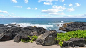 A rocky shoreline with waves crashing against the rocks, a clear blue sky, and a distant horizon.