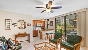 A spacious living area with a sofa, two wicker chairs, a glass coffee table, a kitchen with wooden cabinets, a ceiling fan, and a balcony with a view of a garden.