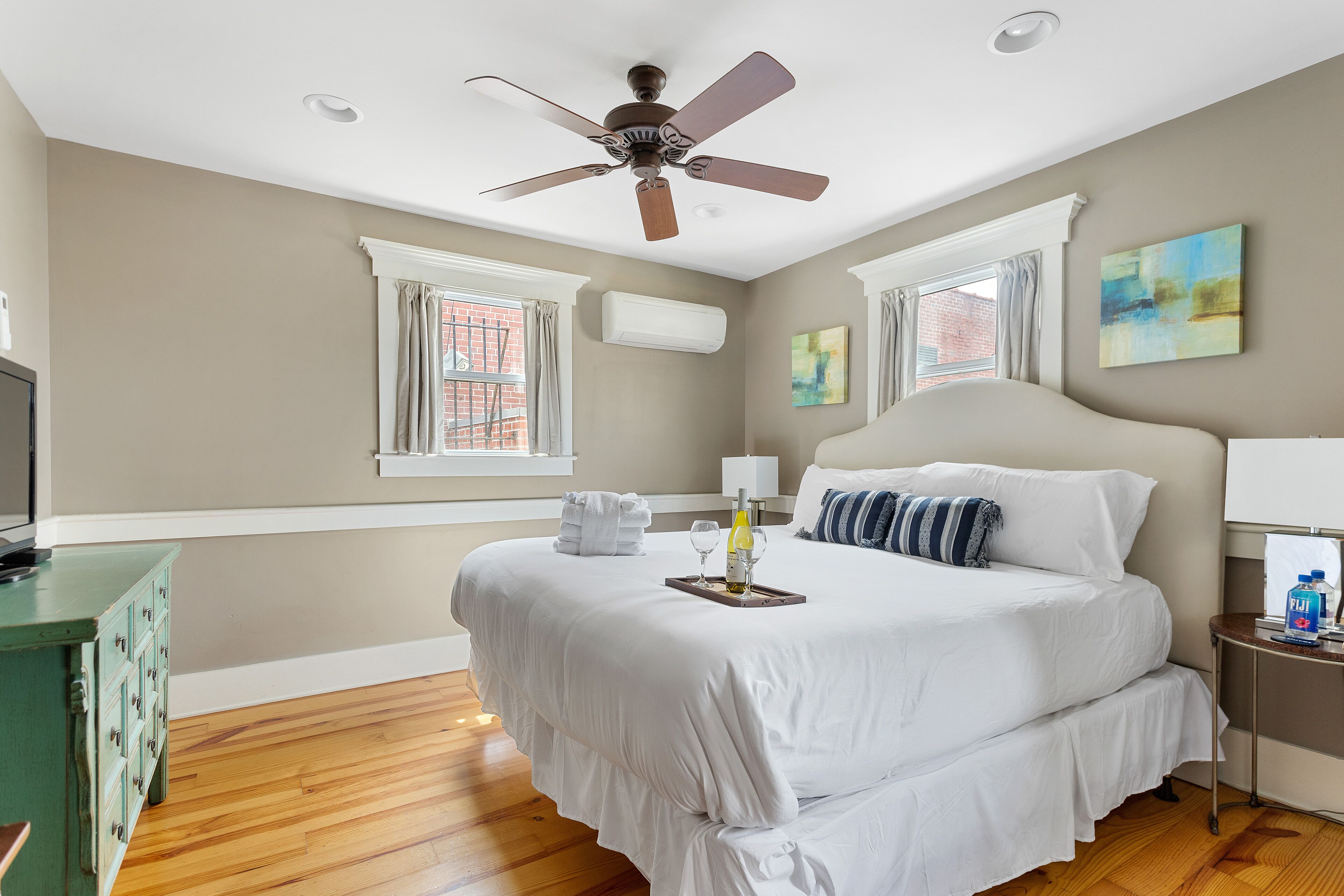 A bedroom with a large bed, a ceiling fan, two windows with curtains, a bedside table with a lamp, and a wall-mounted air conditioner.