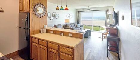 A modern kitchen with wooden cabinets, a central island, and a dining area with a view of the beach.