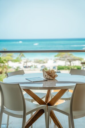 Een ronde tafel met een houten blad, omringd door witte stoelen, met uitzicht op een strand, de oceaan en een heldere lucht.