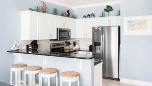 Una cucina con mobili bianchi, un frigorifero in acciaio inox, un forno a microonde e una macchina da caffè. Sono presenti tre sgabelli in vimini e pesci decorativi appesi al muro.