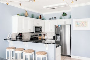 Una cocina con gabinetes blancos, un refrigerador de acero inoxidable, un microondas y una cafetera. Hay tres taburetes de mimbre y peces decorativos en la pared.