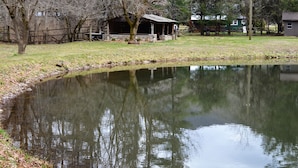 A rustic cabin by a pond with a stone foundation and a wooden roof, surrounded by trees and a fence. - Waterstone Cottage - Outdoor Fireplace, Serene Pond, Rocking Chair Front Porch (Canton)