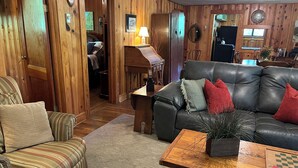 A cozy living room with a leather sofa, striped armchair, wooden walls, and a small table with a chessboard. - Waterstone Cottage - Outdoor Fireplace, Serene Pond, Rocking Chair Front Porch (Canton)