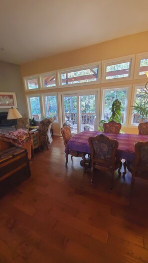 A dining area with a table covered in a purple tablecloth, surrounded by wooden chairs. Large windows with a view of the outdoors are visible.
