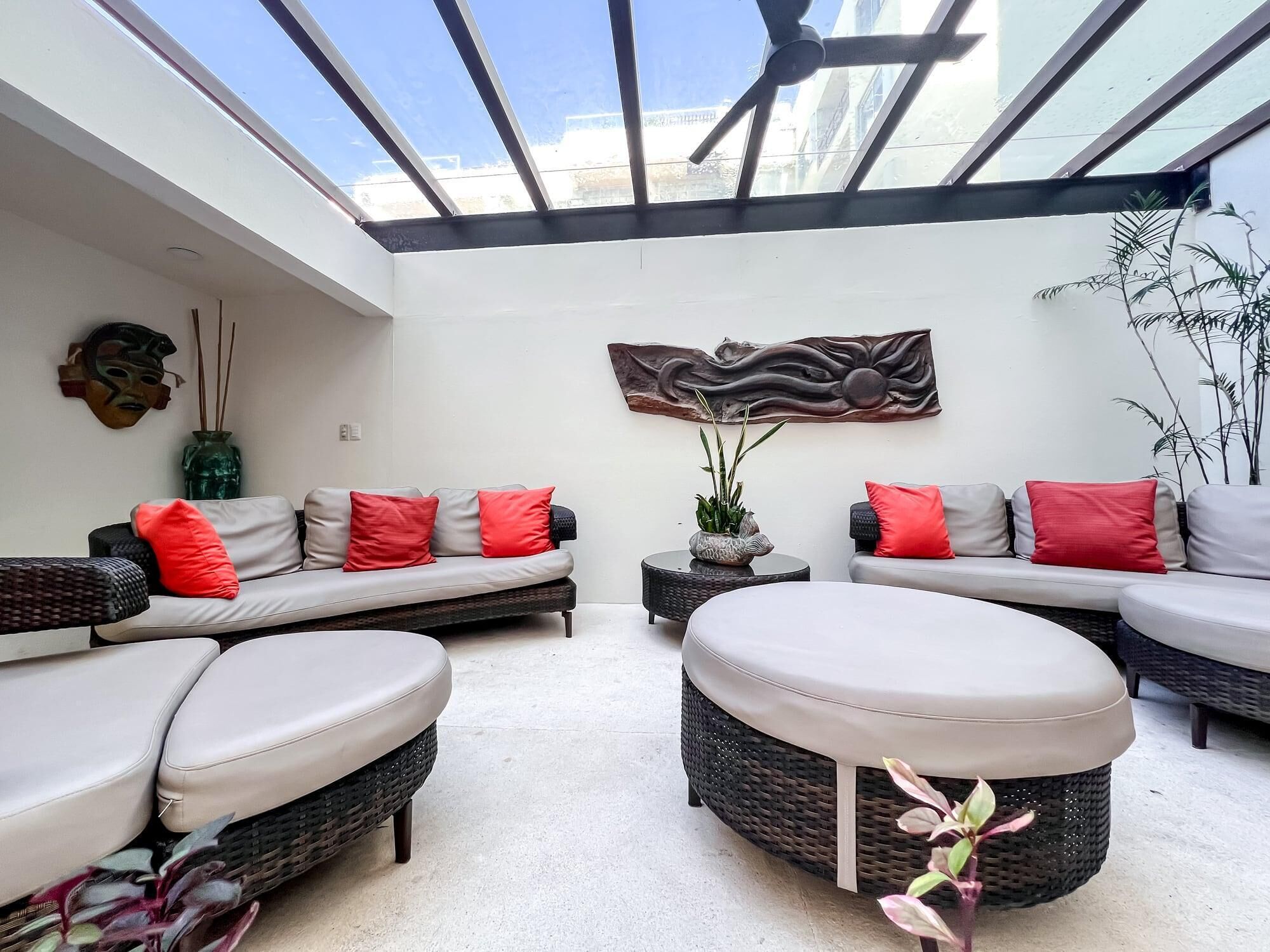A sunlit room with a glass ceiling, a sofa set with red and grey cushions, a round wicker ottoman, and a wall-mounted abstract art piece.