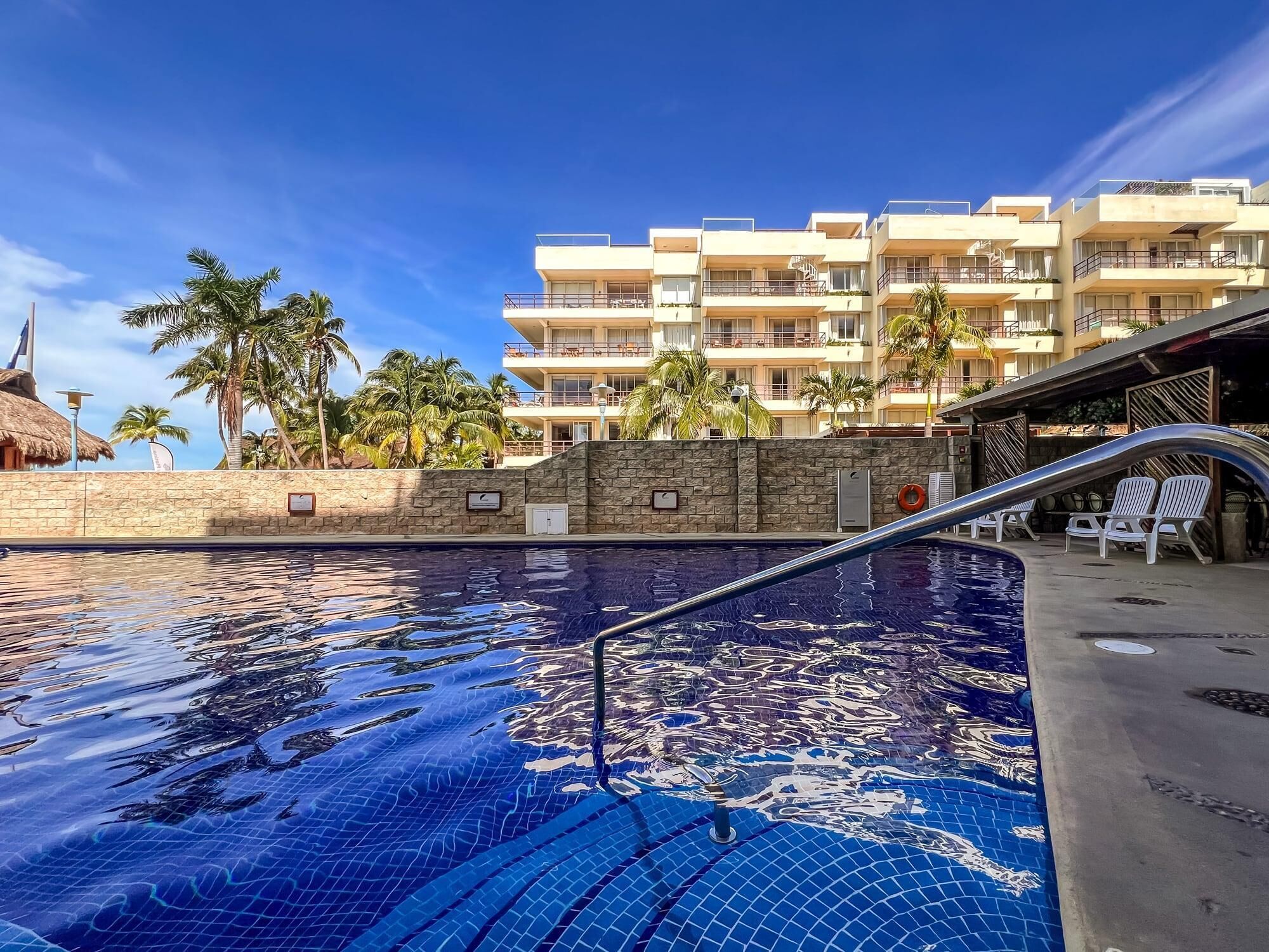 A swimming pool with a clear blue mosaic design, surrounded by a stone wall and a building with balconies and palm trees.