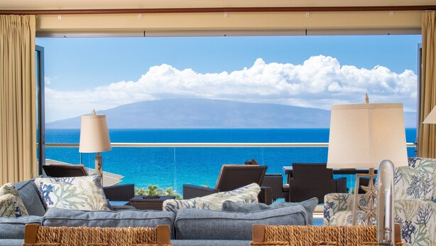 A balcony with a view of the ocean and a mountain, furnished with a sofa, chairs, and lamps.