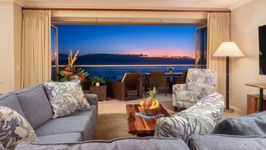 A modern living room with a view of the sea at sunset, featuring a sofa, armchairs, a coffee table with fruit, and a dining area.