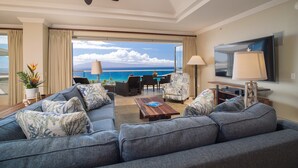 A spacious living room with a large gray sofa, a wooden coffee table, and a ceiling fan. A large flat-screen TV is mounted on the wall, and there is a view of the ocean through the glass doors.