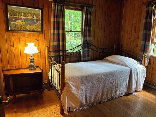 A wooden room with a bed, a nightstand with a lamp, a painting on the wall, and a window with curtains. - Beautiful Mountain Lake Cabin (Piermont)