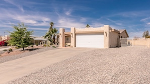 A single-story house with a garage, surrounded by a gravel driveway and a small garden with palm trees and shrubs. - Incredibly Spacious Floorplan with Pool- 3 Bedroom/2 Bath Home (Lake Havasu City)