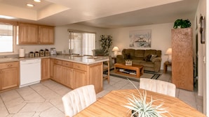 A modern kitchen with wooden cabinets, a dishwasher, and a dining area with a wooden table and chairs. There is a living area with a sofa and a coffee table. - Incredibly Spacious Floorplan with Pool- 3 Bedroom/2 Bath Home (Lake Havasu City)