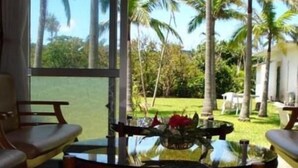 Una habitación con un ventanal grande, una mesa de vidrio con un florero y vistas a un jardín tropical con palmeras.