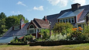 Uma casa grande de dois andares com telhado inclinado, base de pedra e um jardim bem cuidado com diversas plantas e flores.