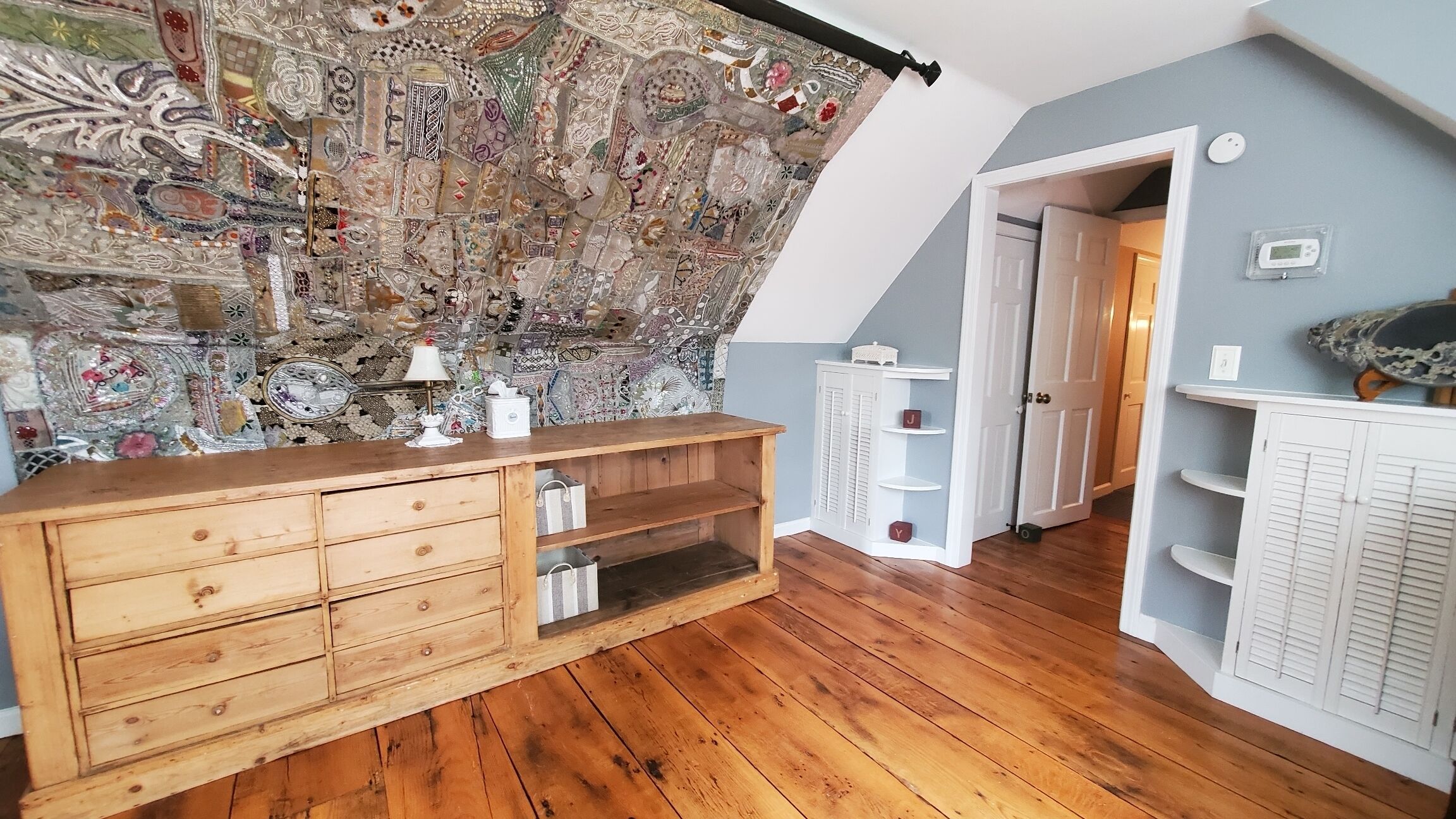 A room with a wooden dresser, a patterned tapestry on the wall, and a white cabinet with shelves.