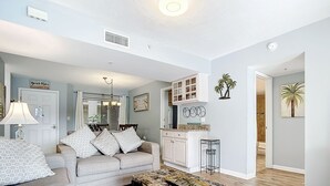 Un salón con pared azul claro, un sofá color beige con cojines estampados, una mesa de centro con cubierta decorativa y una zona de cocina con armarios blancos y encimera.