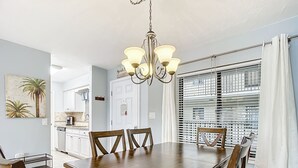 Un comedor con mesa y sillas de madera, una araña y vistas a través de una ventana con persianas.