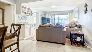 Una sala moderna con un sofá, una mesa de centro y un televisor. Hay un comedor con una mesa y sillas de madera. La habitación tiene un ventanal con vistas a edificios.