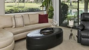 A living room with a beige sofa, a black ottoman, a black leather chair, and a large window offering a view of a park. - Fantastic Mt Views From this Beautiful Mission Hills Country Club Condo (Rancho Mirage)