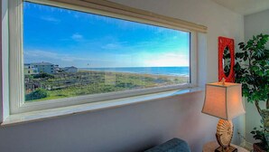 A room with a large window overlooking a beach and a road. There is a lamp with a decorative base on a table.