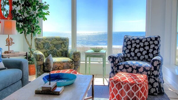 A living room with a view of the ocean, a sofa, a patterned armchair, a small table, and a decorative bowl on a coffee table.