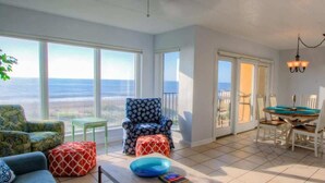 A sunlit room with a view of the ocean, featuring a dining table, chairs, and a patterned armchair.