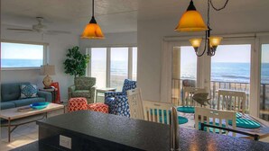 A spacious living area with a large window overlooking the ocean, a dining area with a table and chairs, and a seating area with a sofa and cushions.
