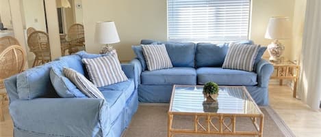 A living room with a blue sofa set, a glass coffee table, wicker chairs, and a large window with blinds.
