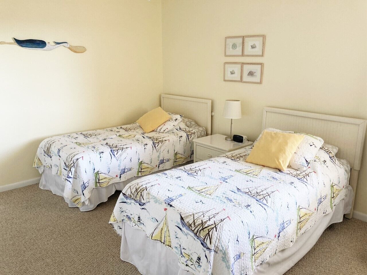 A room with two beds, each with a white bedspread featuring sailboat designs. There are two bedside tables with lamps, a wall-mounted shelf with framed pictures, and a carpeted floor.