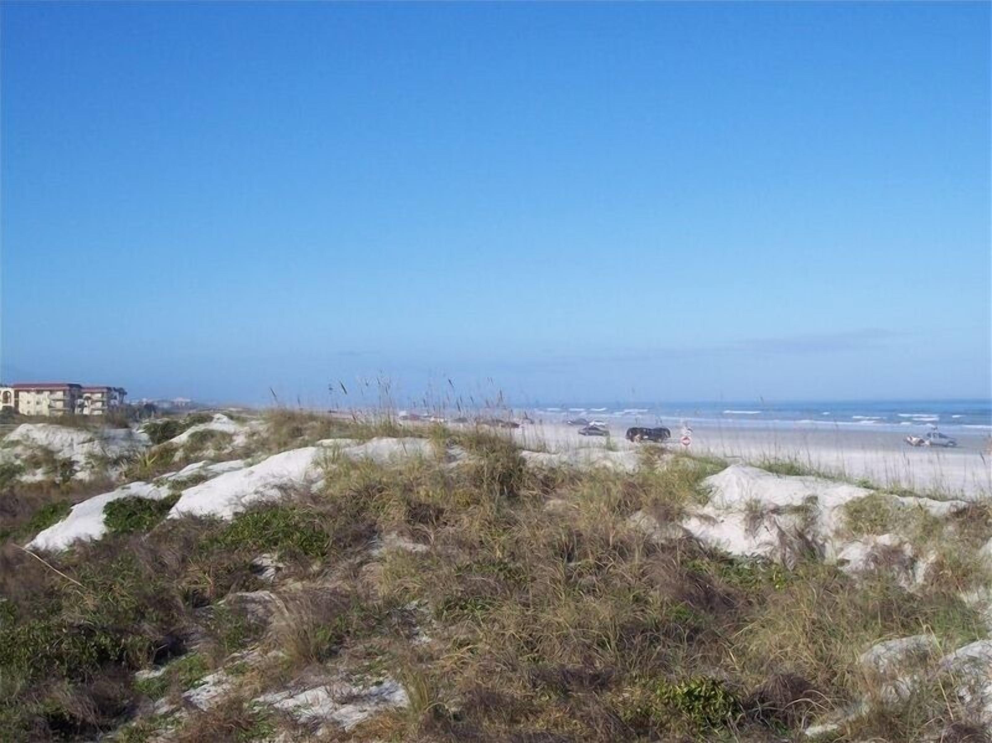 A coastal landscape with dunes, a beach, and distant buildings.