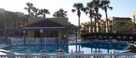 A swimming pool with lounge chairs, surrounded by palm trees and a building with a red roof.