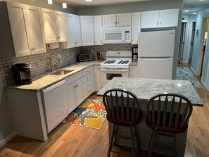 Eine moderne Küche mit weißen Schränken, einer Granit Arbeitsplatte, einem Gasherd, einer Mikrowelle und einem Kühlschrank. Zwei Barhocker und ein farbenfroher Teppich sind ebenfalls vorhanden.