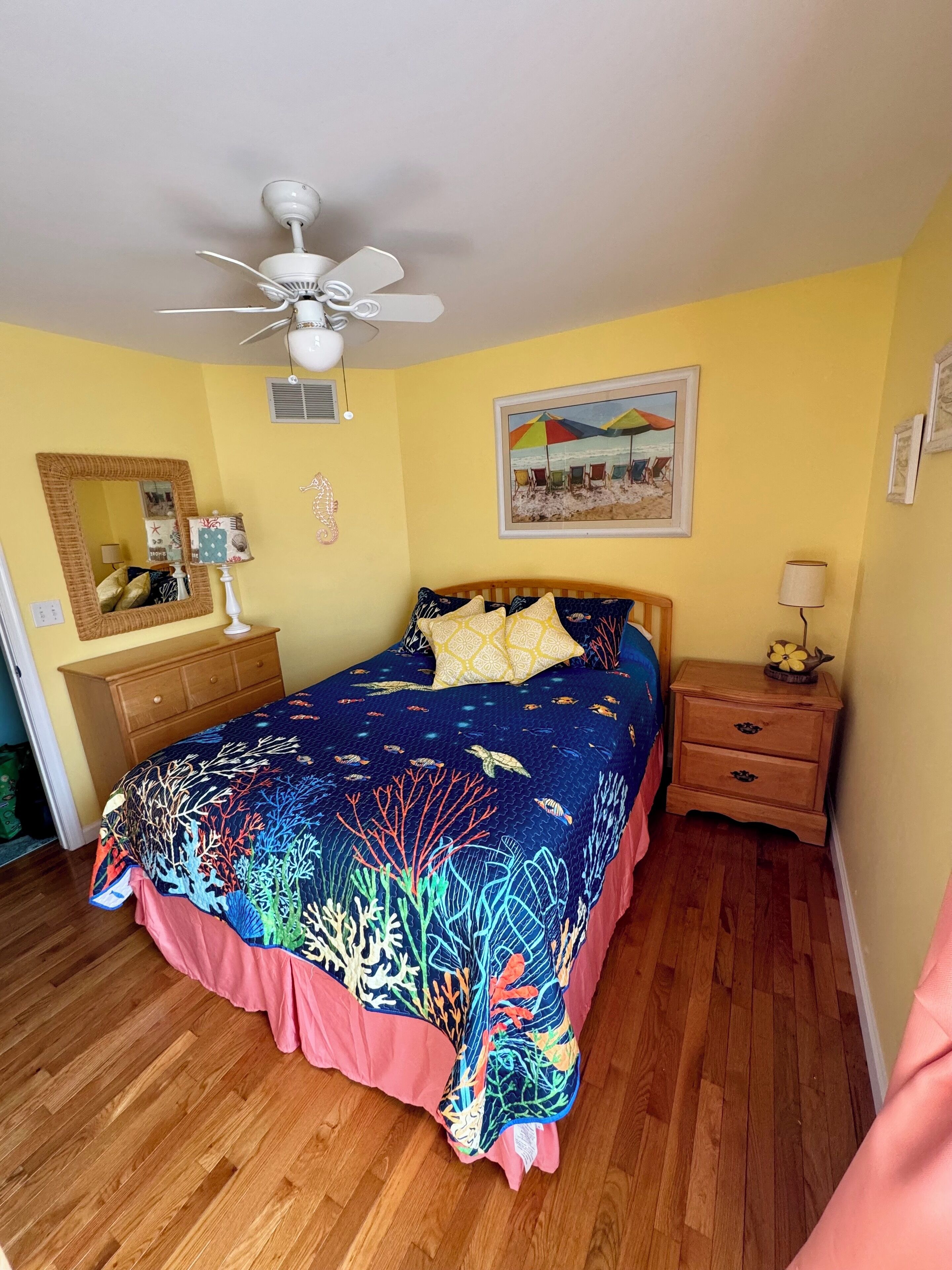 A bedroom with a bed, a ceiling fan, a nightstand, a dresser, a mirror, and a beach-themed painting.