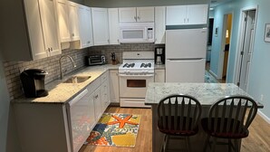 Een moderne keuken met witte kastjes, een magnetron, fornuis en koelkast. Er staat een koffiezetapparaat, een broodrooster en een vloerkleed met een sterren design. Twee stoelen staan bij het keukeneiland.